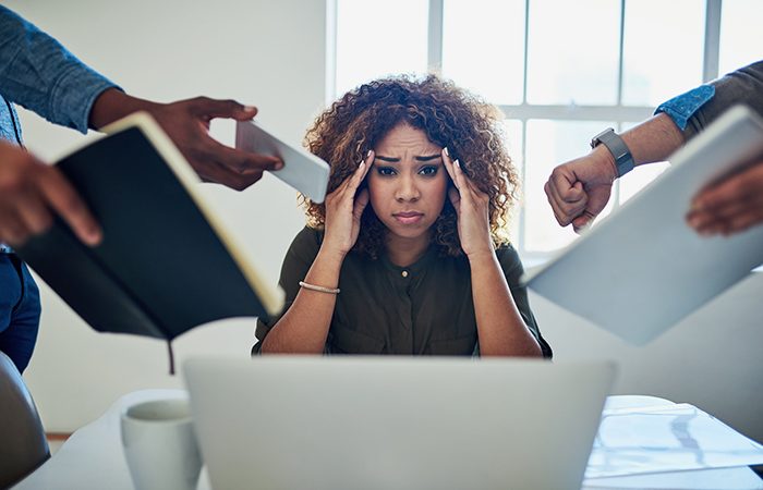 A stressed, African American woman sits at a laptop. employers need to reduce the risk of employee burnout.