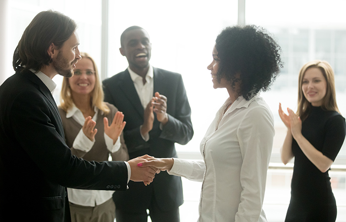 A boss shakes the hand of a woman while other employees applaud, showing how employee engagement is connected to recognition.